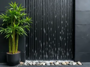 Zen Water Wall - Close-up of a minimalist black slate wall with thin water sheets flowing smoothly down its surface, flanked by potted bamboo and river rocks at the base