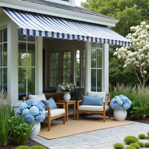 Coastal Hamptons Sunspace - An elegant white-trimmed glass enclosure with navy and white striped awnings, featuring weathered teak furniture, sisal rugs, and blue hydrangeas in white ceramic planters. Coastal grasses and white flowering dogwood trees frame the space