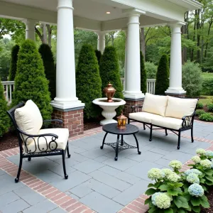 Classic Colonial Veranda - Bluestone patio with traditional brick border, classic white columns supporting overhead structure. Black wrought iron furniture with cream cushions, surrounded by formal boxwood hedges and hydrangea bushes. Copper lanterns and traditional fountain.