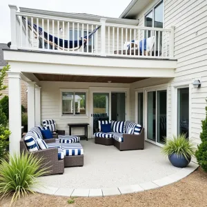 Coastal Cool Escape - Light-colored exposed aggregate concrete patio under a white composite deck. Navy and white striped outdoor furniture, rope hammock, and coastal-themed decor. Large ceramic planters with Maiden Grass and Blue Star Juniper