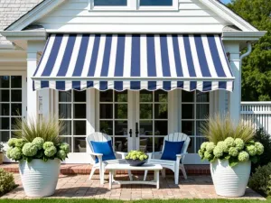 Coastal Canvas Stripe Awning - A classic navy and white striped retractable canvas awning extending over a whitewashed brick patio. Coastal-style furniture with blue cushions sits beneath, surrounded by white ceramic planters filled with coastal grasses and hydrangeas.