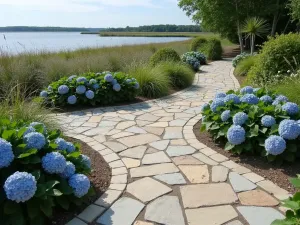 Coastal Paver Design - Light-colored tumbled pavers in a random pattern, reminiscent of beach stones, surrounded by coastal grasses and blue hydrangeas, overlooking water